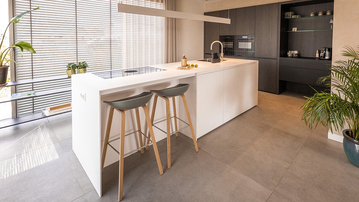 Corian kookeiland met bar en strakke keuken kastenwand met apparatuur. Corian kookeiland met bar en strakke keuken kastenwand met apparatuur. Luxe keuken, strakke keuken, minimalistisch design van B Dutch in Cuijk. Met solid surface Corian kookeiland, keukeneiland, en strakke houten keuken kastenwand. Boven het keukeneiland hangt de B Dutch lineair smalle hanglamp met led verlichting.
